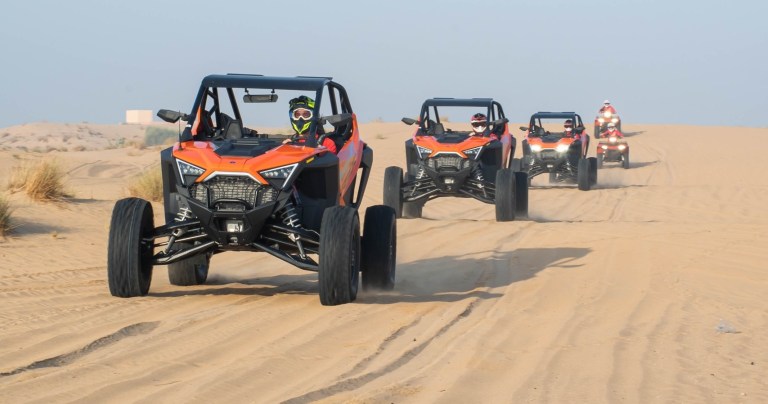 Four off-road vehicles driving on a sandy desert trail.