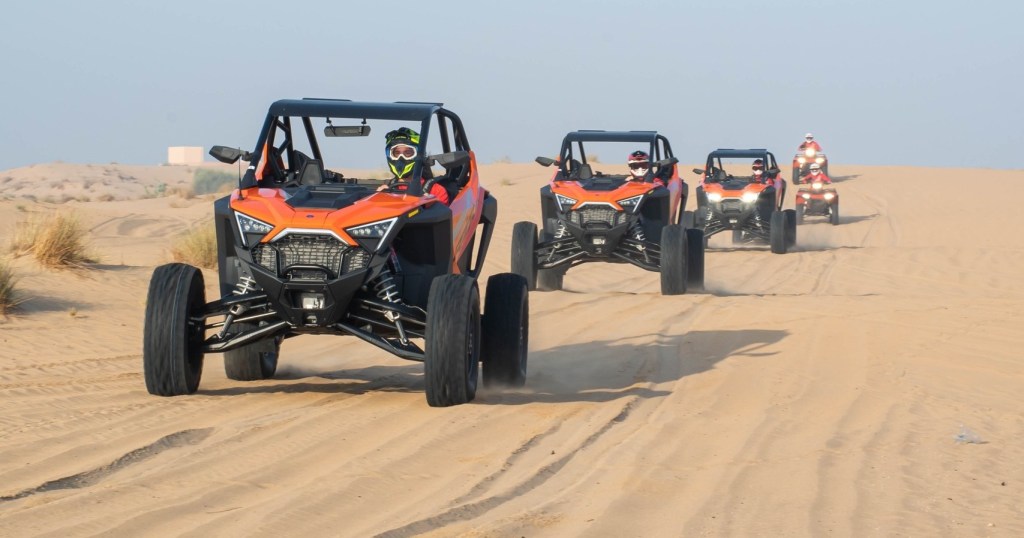 Four off-road vehicles driving on a sandy desert trail.