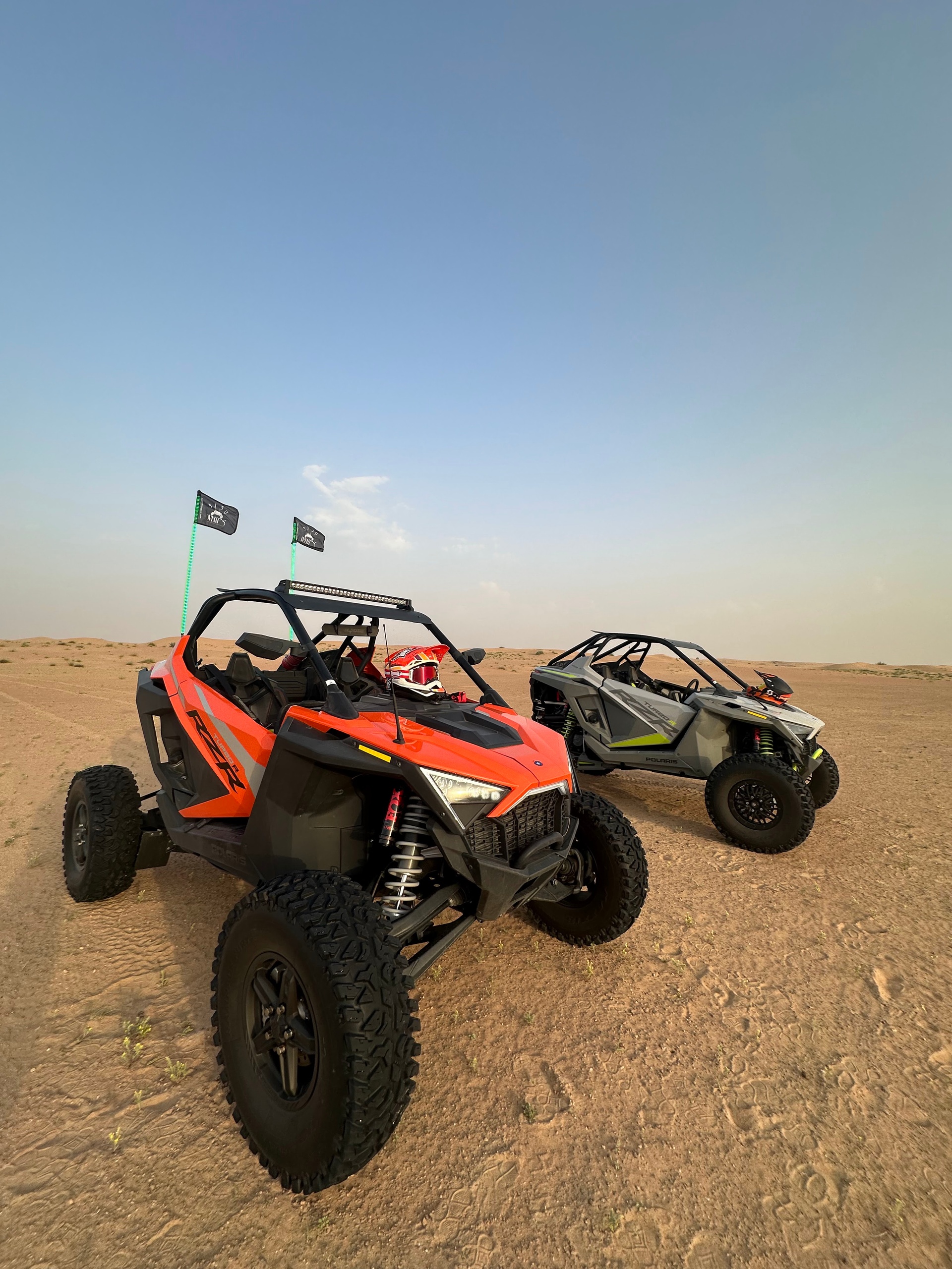 Two off-road vehicles on desert terrain with flags, clear sky.