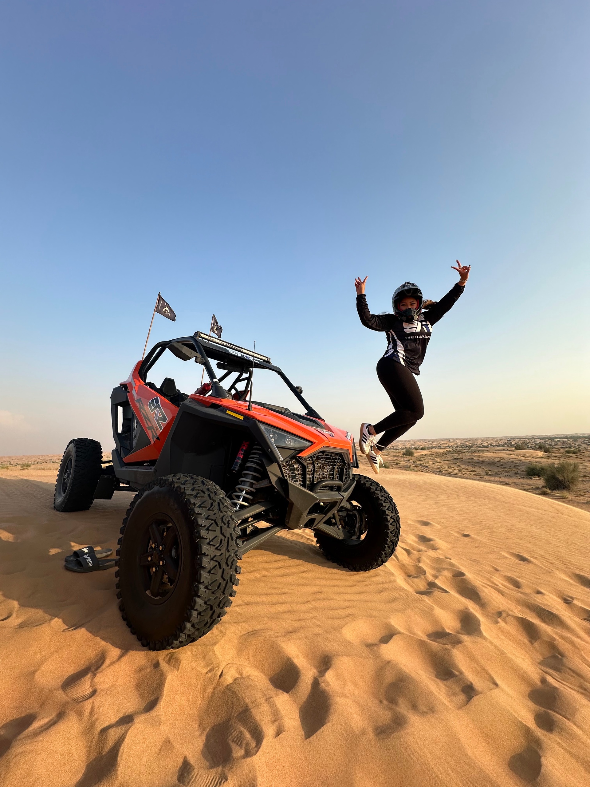 Person jumping near an off-road vehicle on sandy dunes under a clear sky.