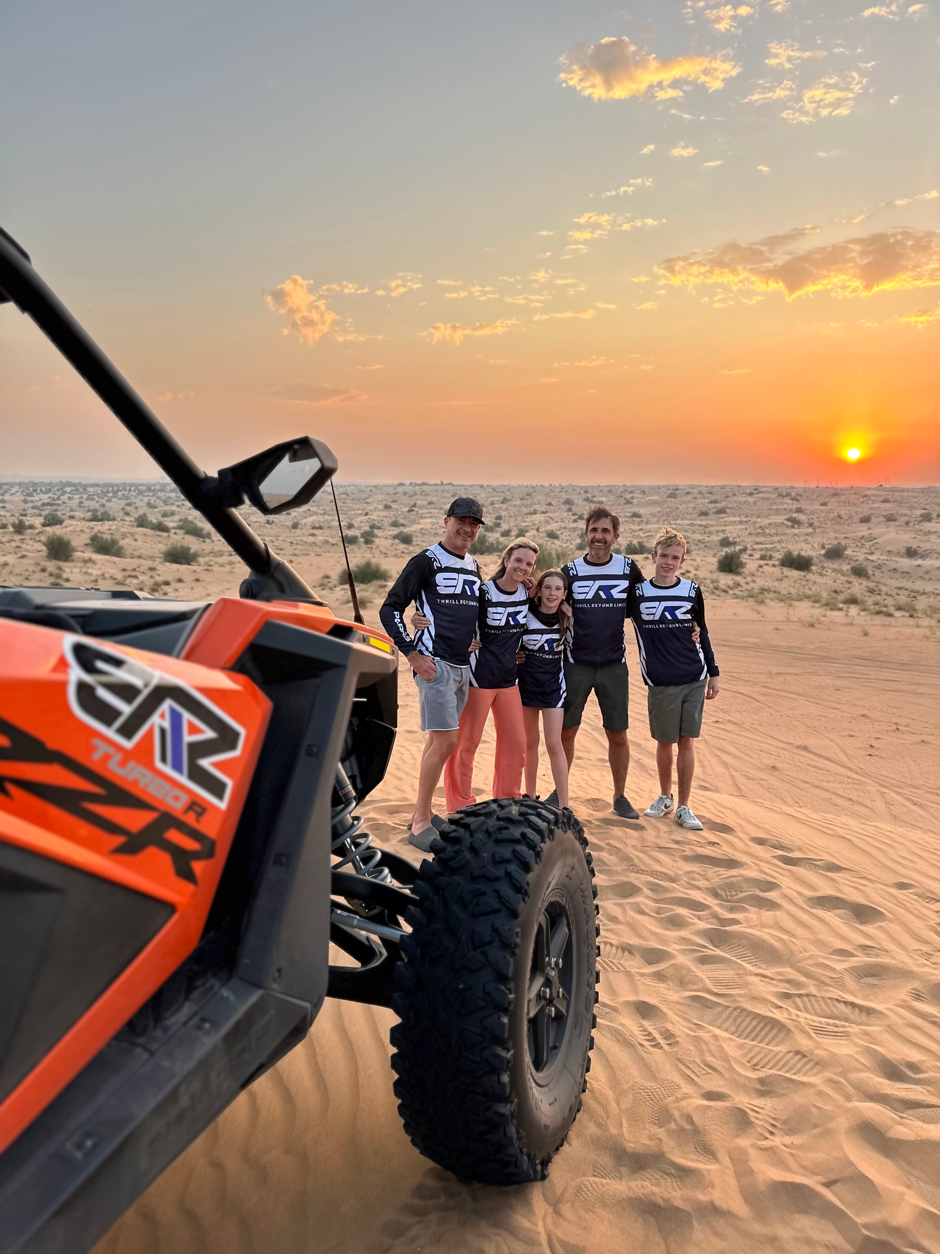 Group by dune buggy on sandy desert, sunset in background.