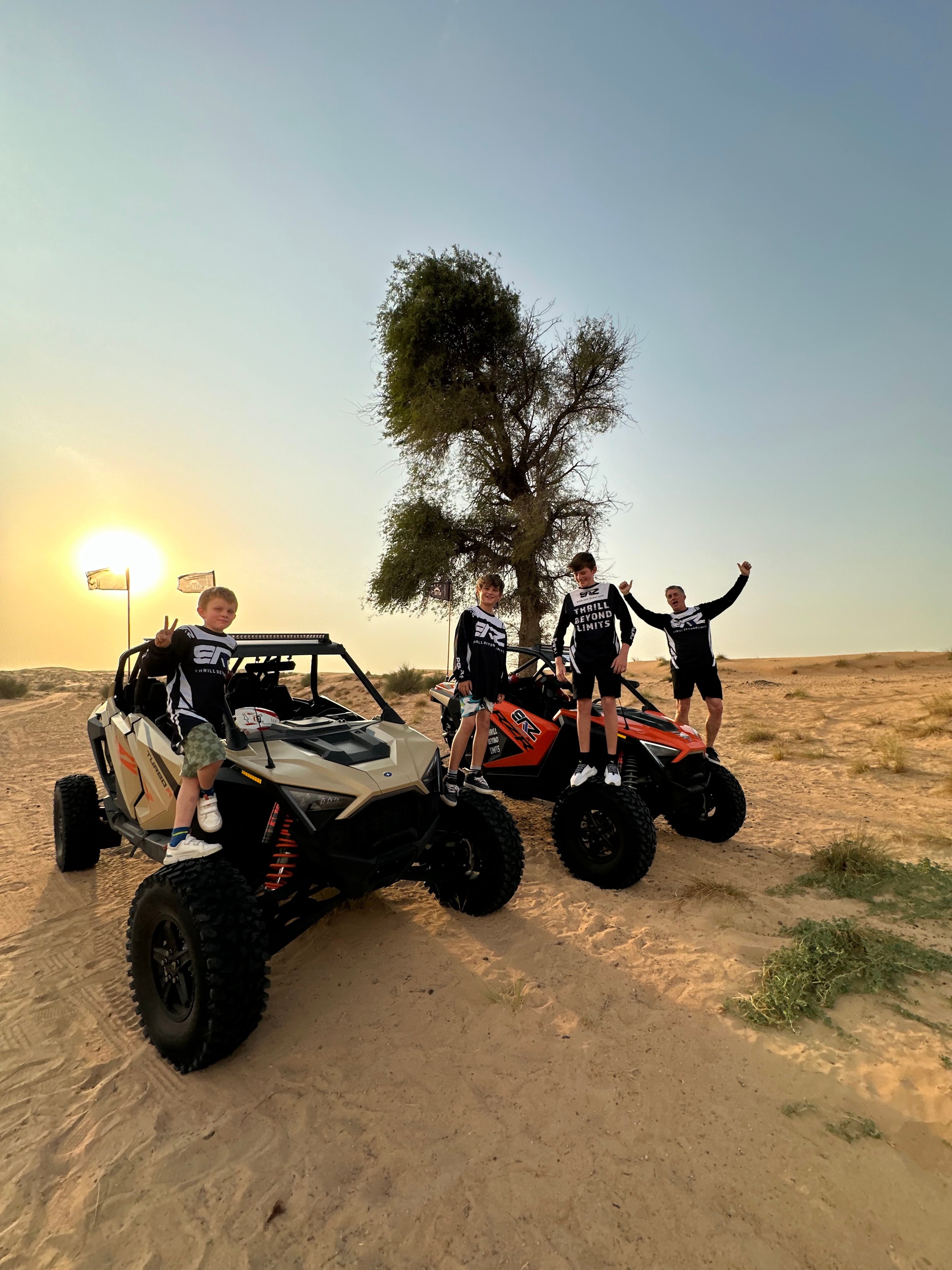 Four people in black outfits pose on two dune buggies in a sandy desert with a sunset.