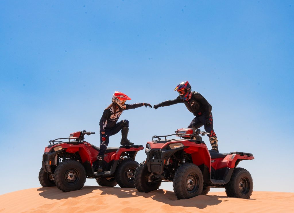 Two people on ATVs high-fiving on a sandy dune under a clear blue sky.