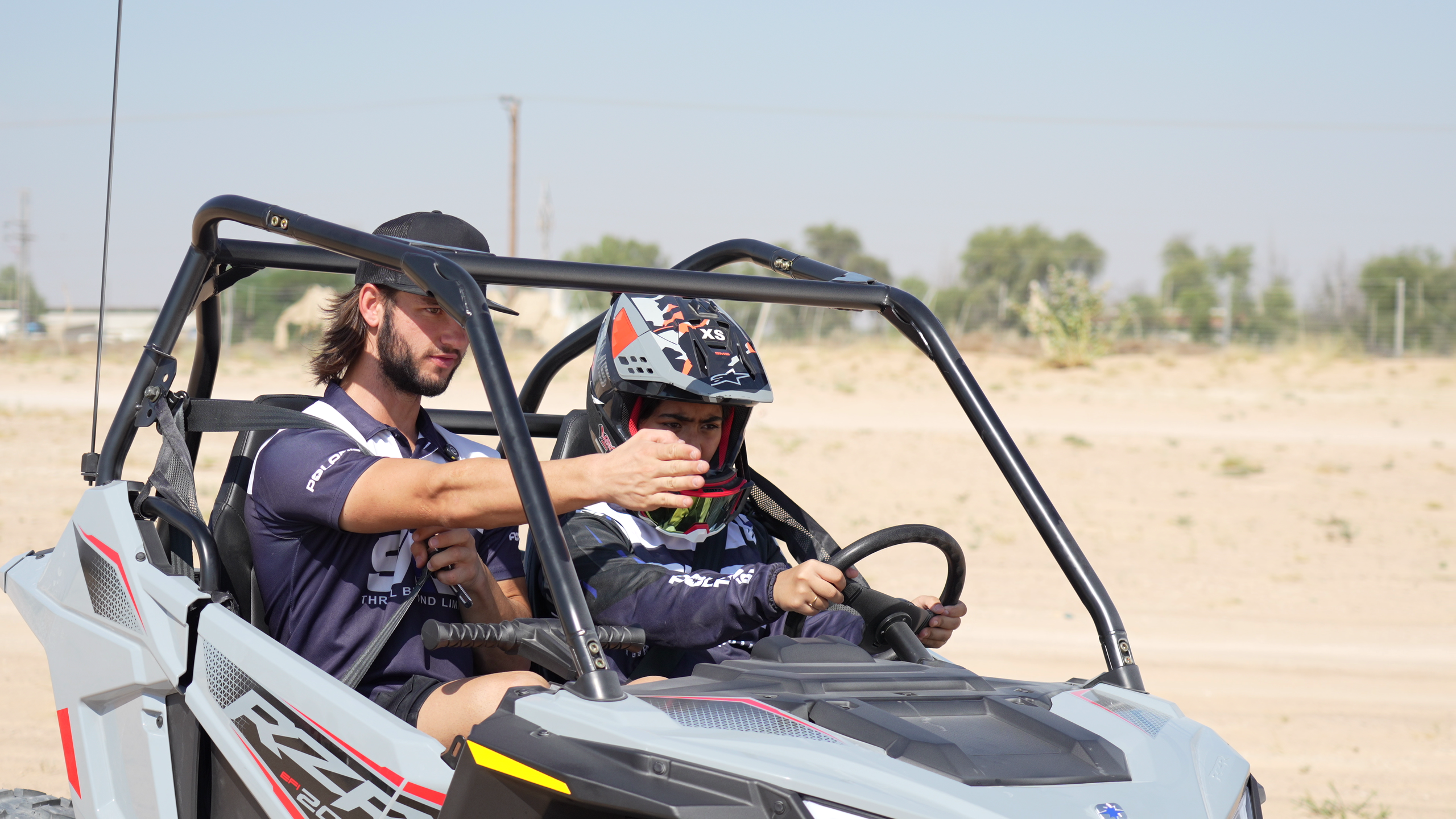 Two people in a dune buggy, one driving, on a sandy terrain.