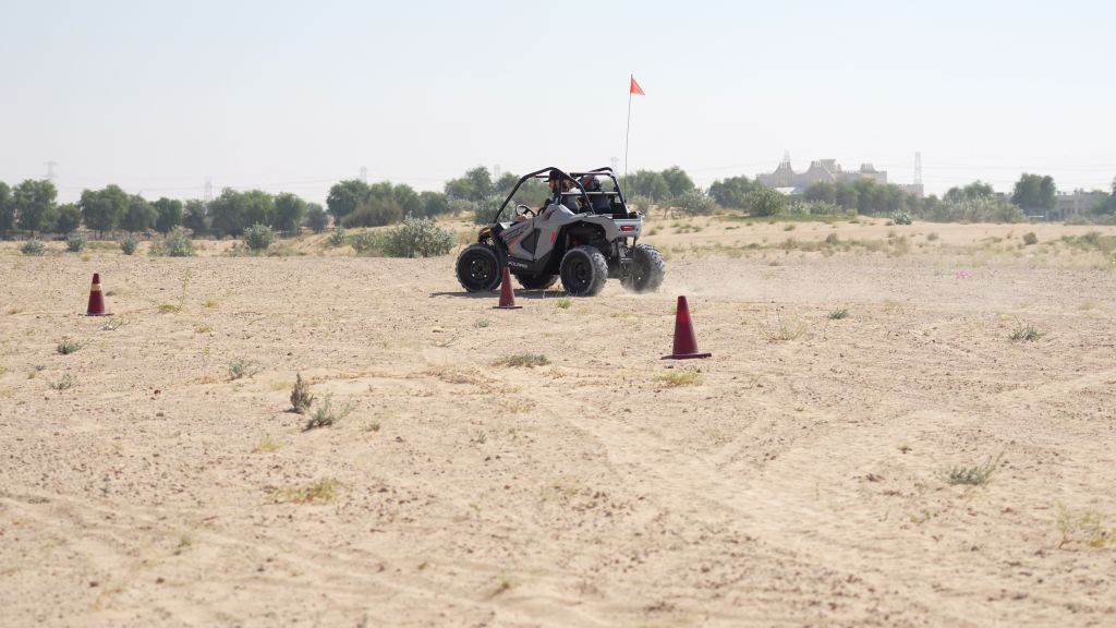 Off-road buggy driving on desert terrain with cones and a distant tree line.