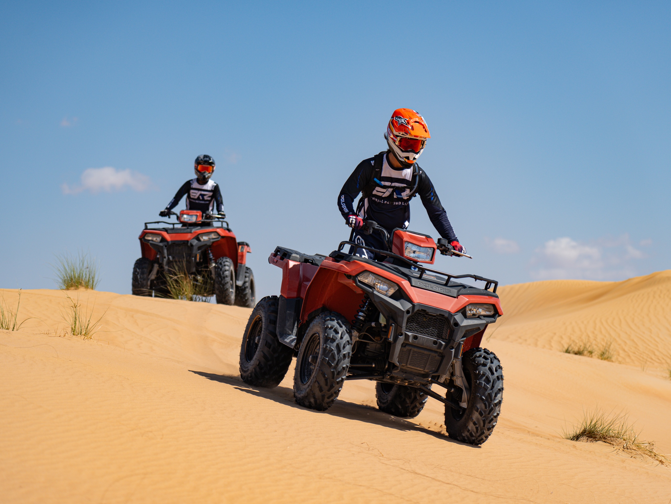 Two people riding red ATVs on sand dunes under a blue sky.