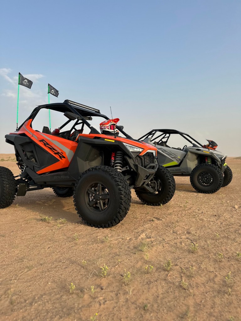 Two off-road buggies on sandy terrain with flags in the background.