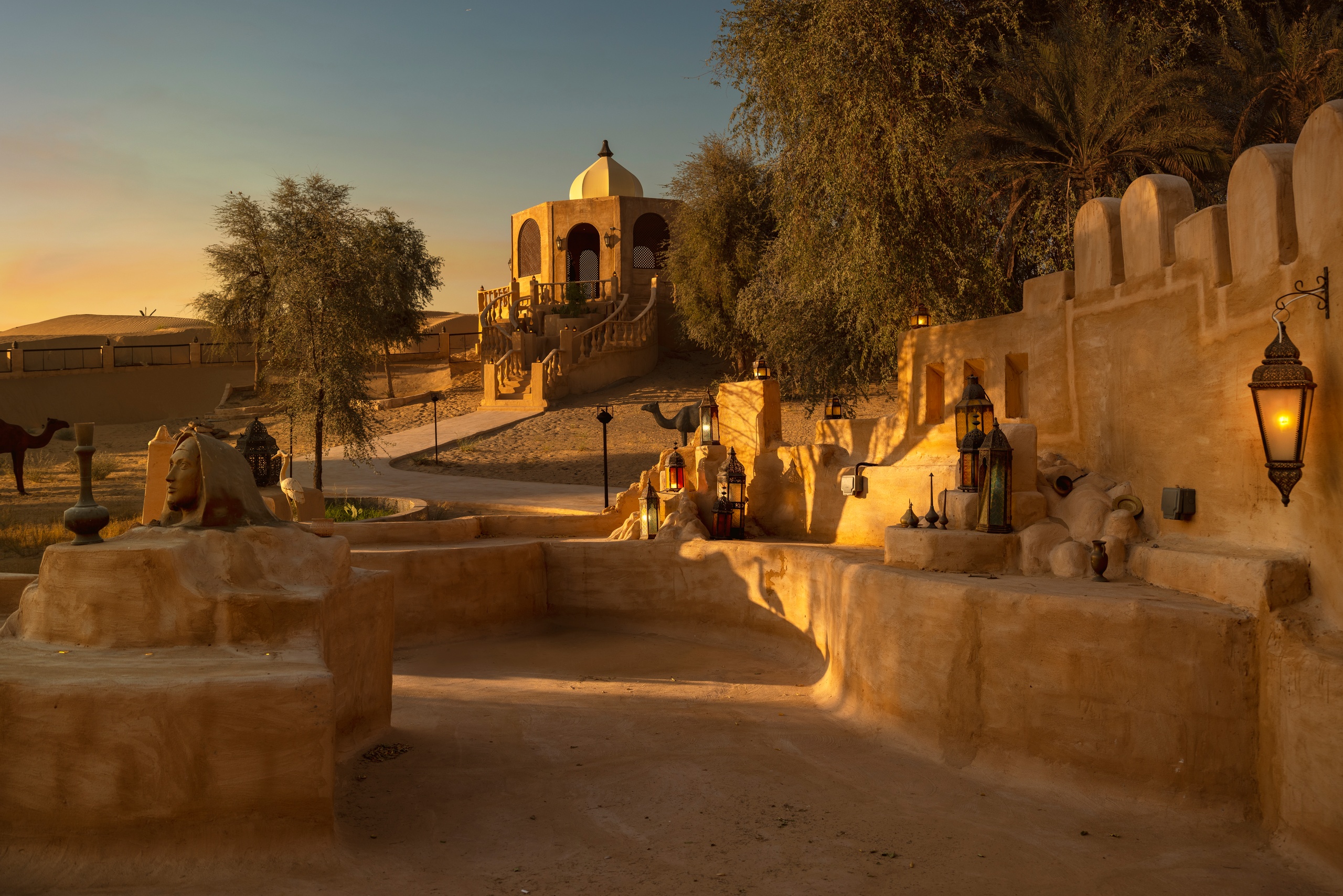 Desert scene with stone structures, lanterns, and a domed pavilion at sunset.