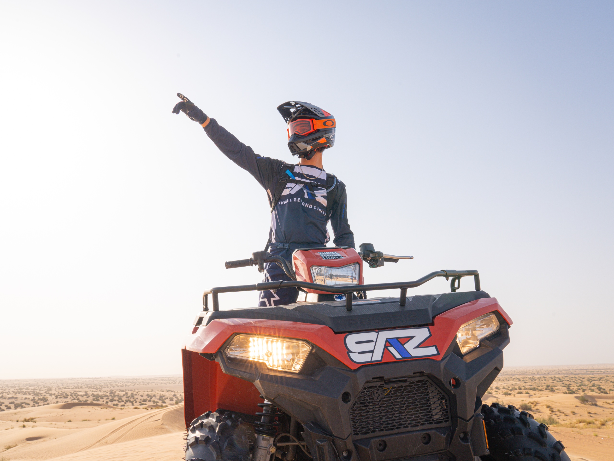 Person on ATV in desert, pointing forward, wearing helmet and goggles.