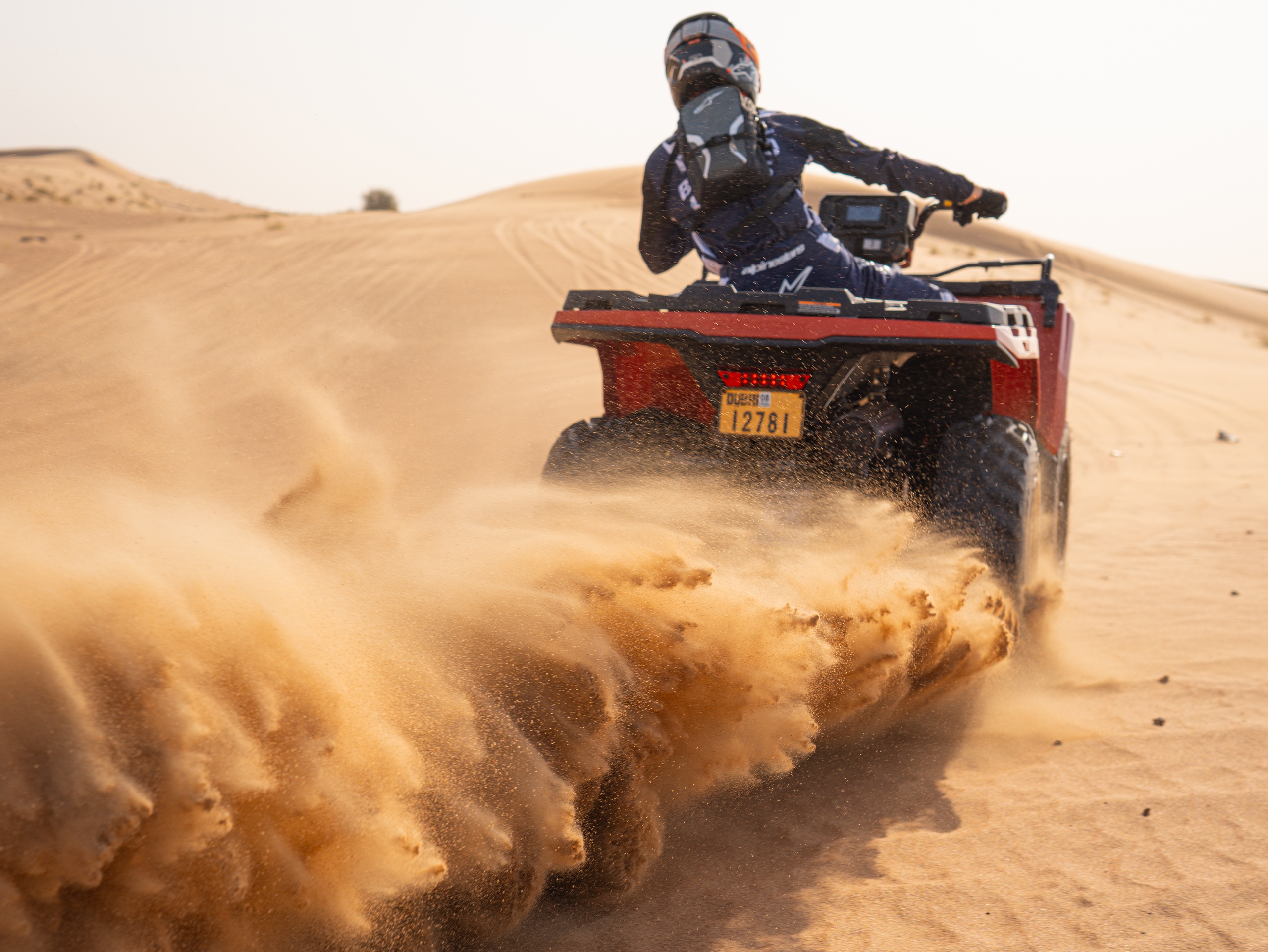 Person riding a quad bike through desert sand, kicking up dirt.
