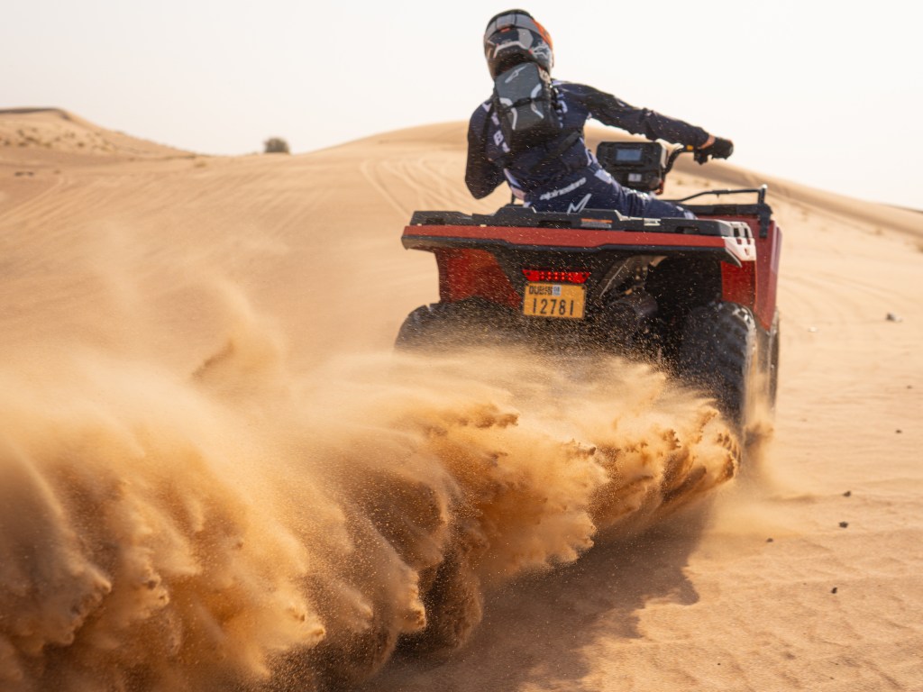 Person riding a quad bike through desert sand, kicking up dirt.