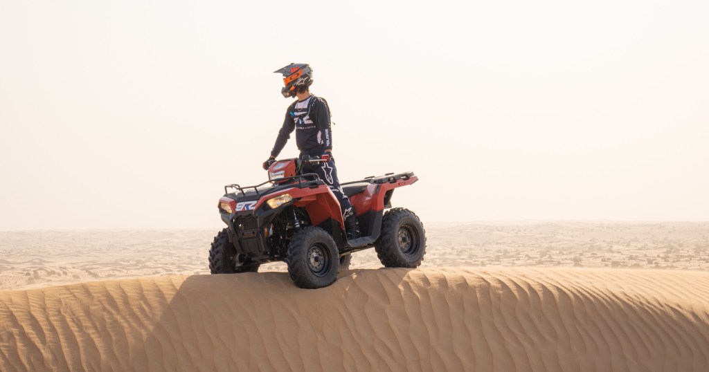 Person on red ATV standing on sandy desert ridge.