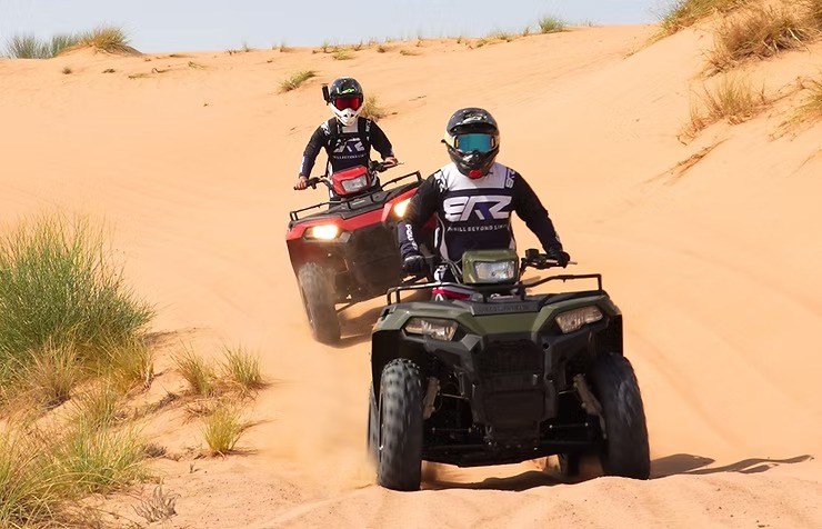 Two people riding ATVs on sandy dunes with grass patches.