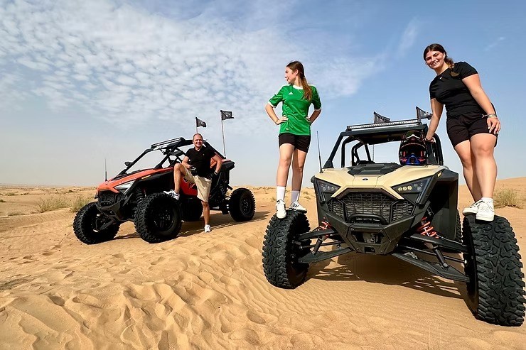 Three people pose with two dune buggies on desert sand.