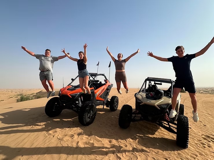 Four people jumping beside dune buggies in a desert setting under blue sky.