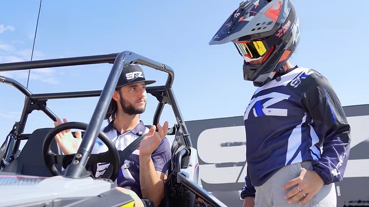 Two people in racing gear, one sitting in a UTV, the other standing with helmet, on a sunny day.