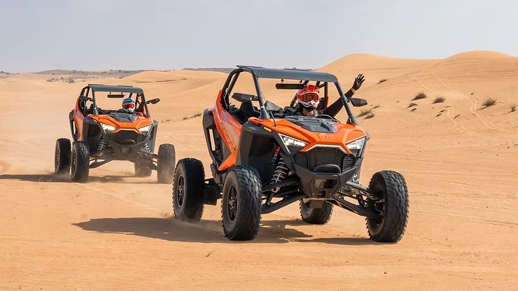 Two orange dune buggies driving in sandy desert with riders in helmets.