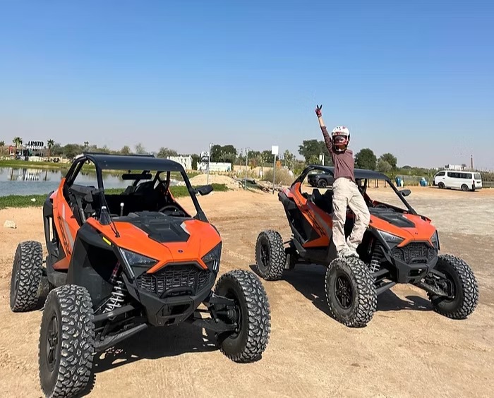 Person in helmet stands on one of two parked orange dune buggies, raising a hand in a peace sign.