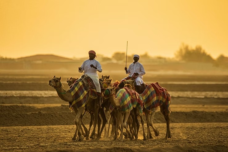 Two men in traditional attire ride camels at sunset on a sandy landscape.