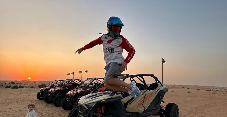 Person jumps off dune buggy in desert at sunset; child nearby.