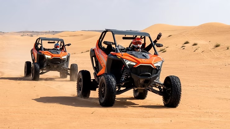 Two orange dune buggies driving on desert sand with raised dust.