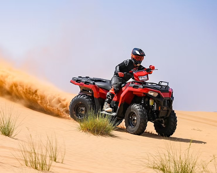 Person riding red ATV, kicking up sand on a desert dune.