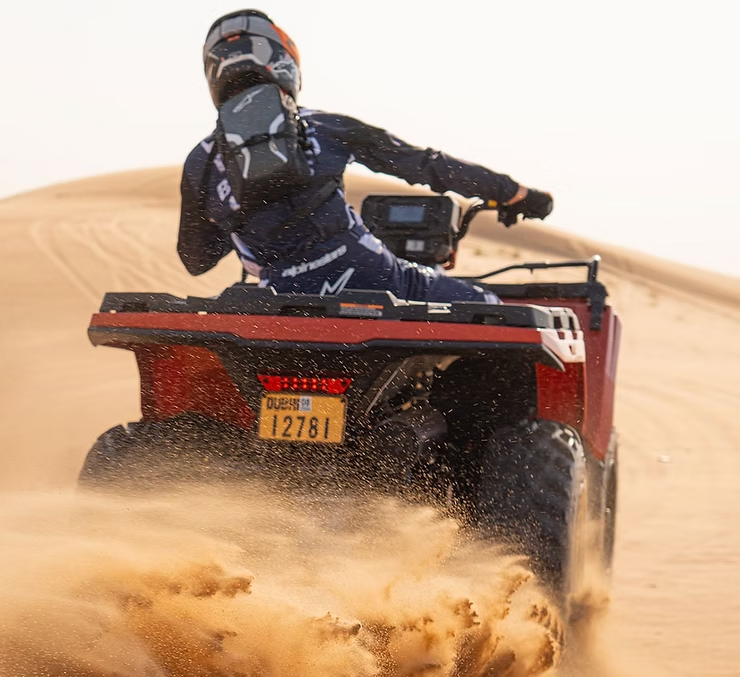 Person riding an ATV on sand dunes, sand flying, wearing helmet and gear.