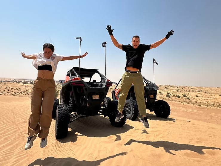 Two people jump in front of desert vehicles on sandy terrain.